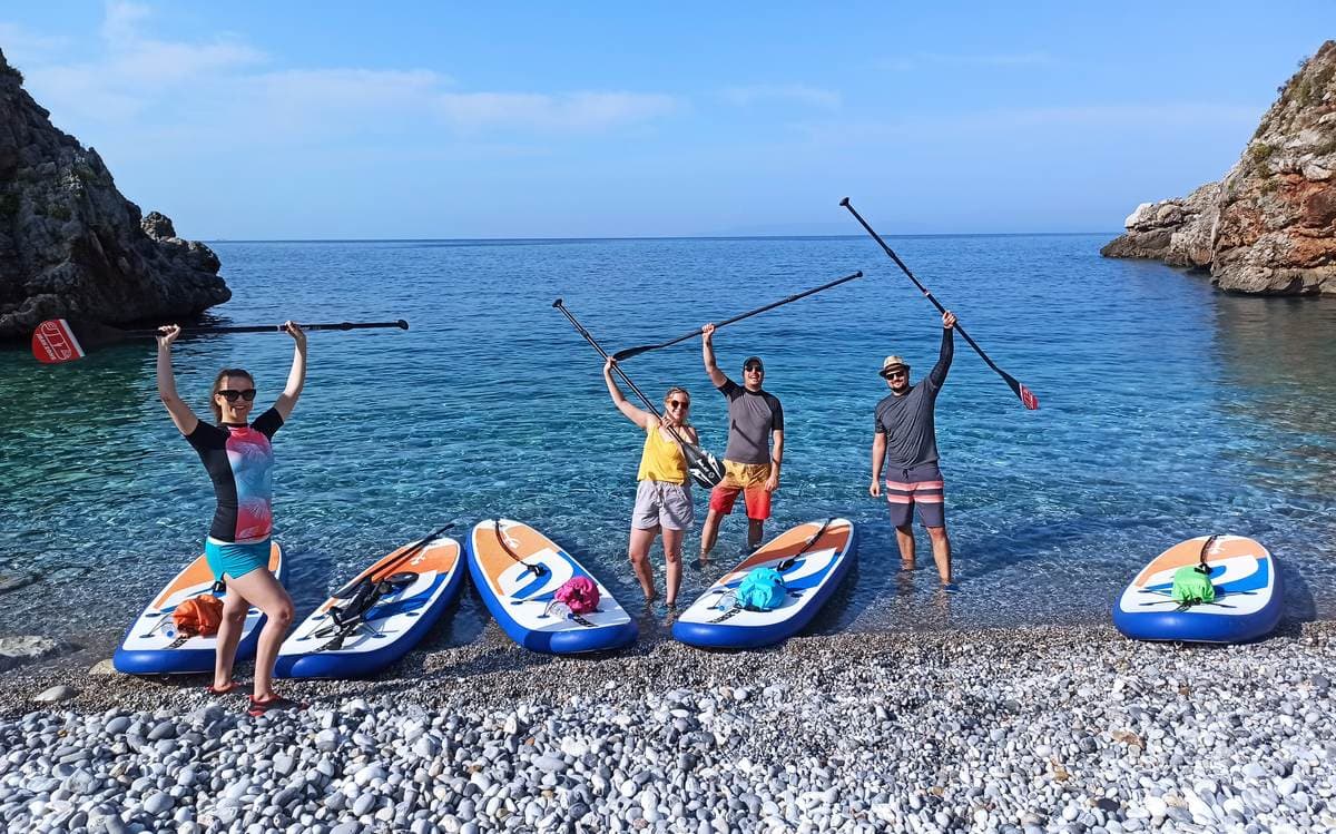 A guided group during one of our paddleboarding tours in Greece posing with SUP boards on a pebble beach.