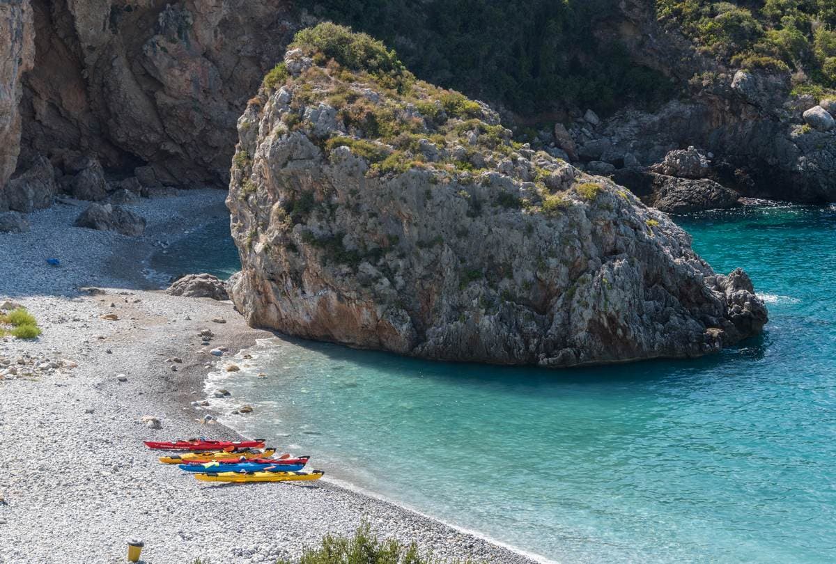 Brightly colored kayaks parked on a white pebble beach next to a giant sea stack during a sea kayak Kardamyli trip.