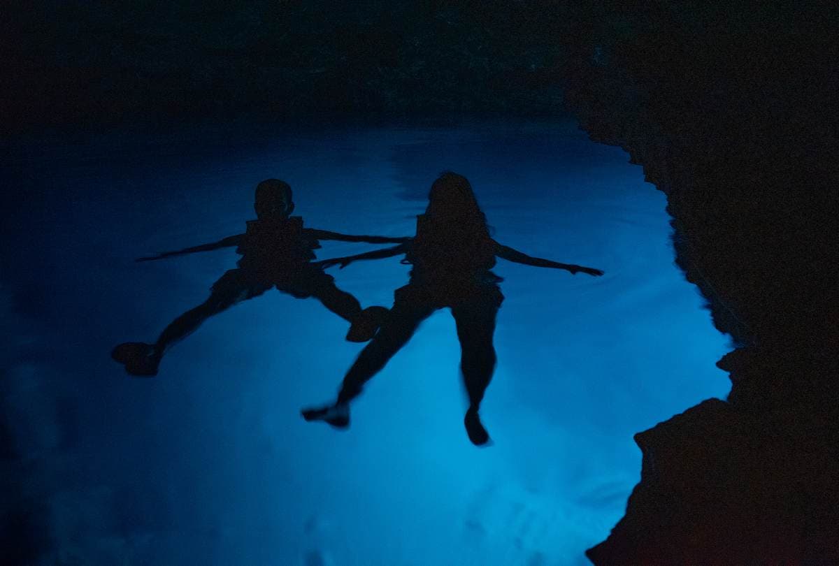 Silhouette of two people swimming in the glowing neon-blue water of a sea cave in Mani, Greece.
