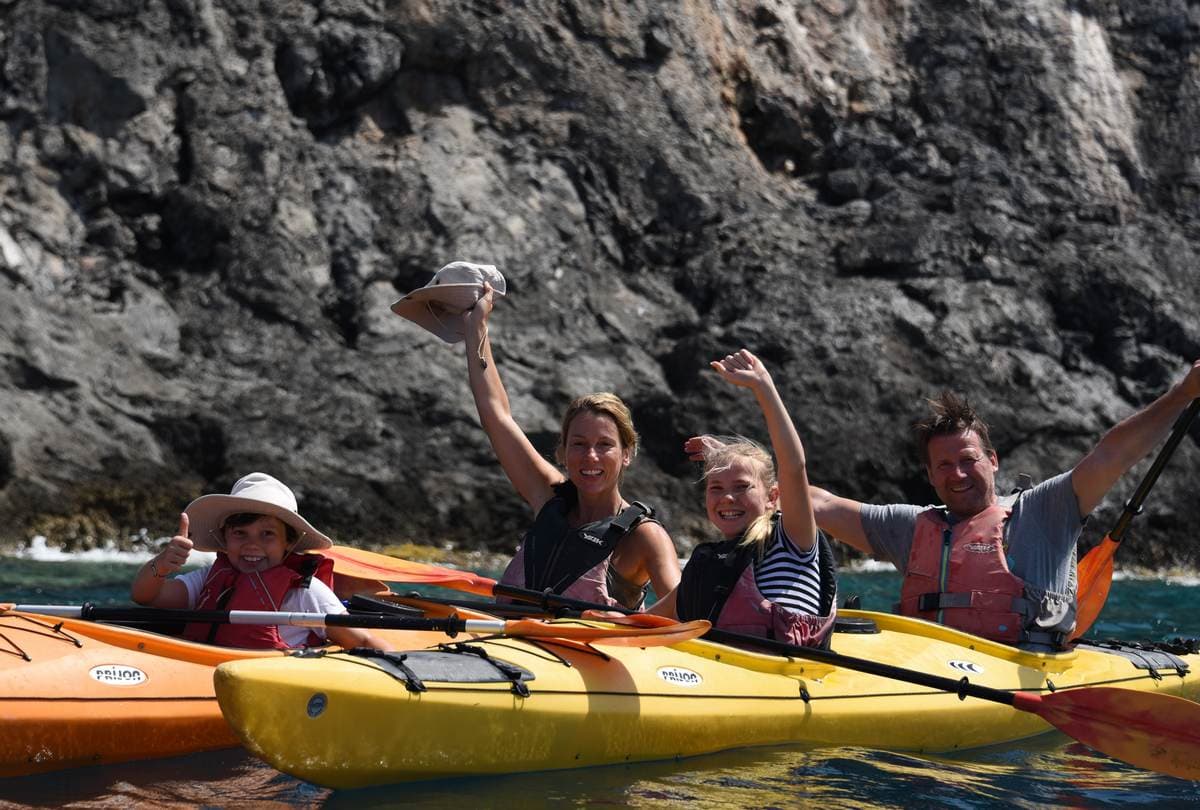 A happy family of four waving and smiling from their kayaks in front of a dark rock cliff.