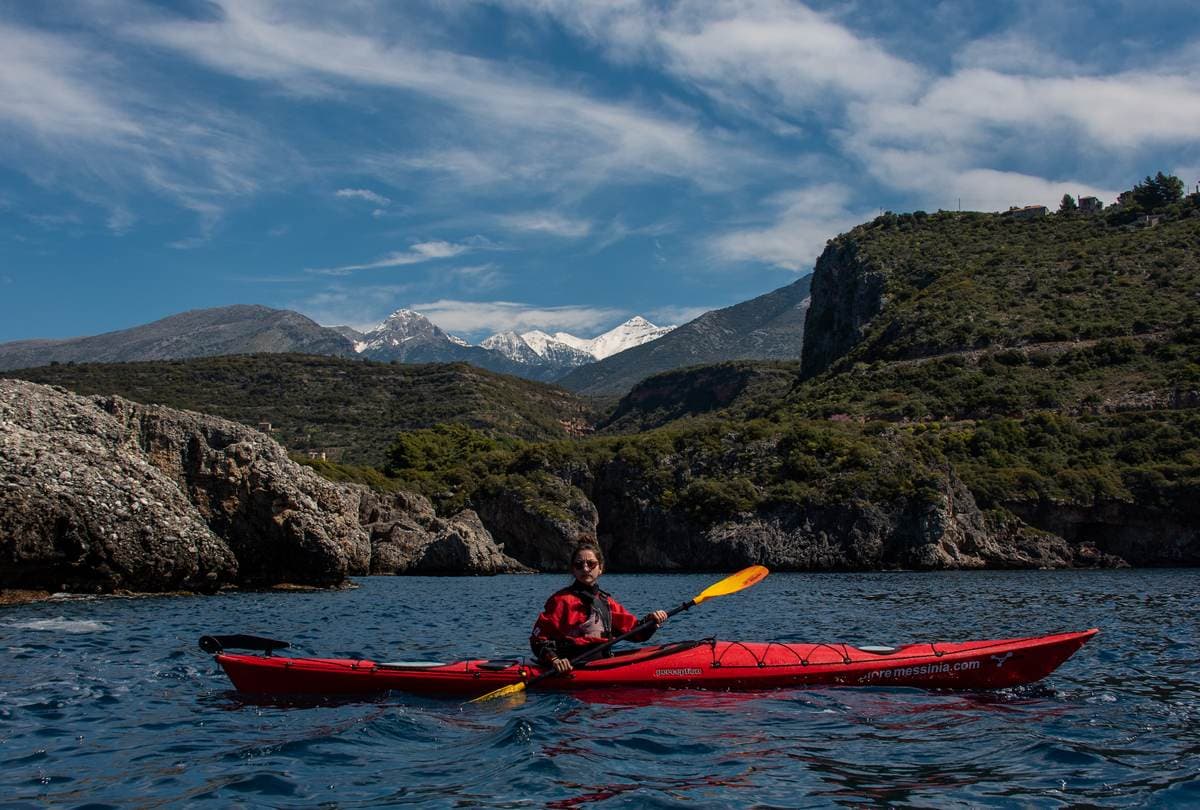 A woman in a red sea kayak paddling in the Messinian Gulf with the snow-capped Taygetos mountains in the background.