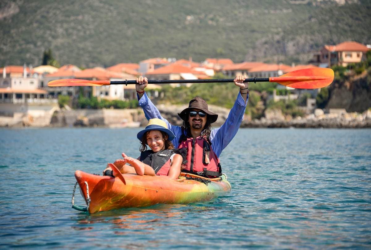 A father and daughter smiling together in an orange tandem kayak on the calm blue waters of Kardamyli.