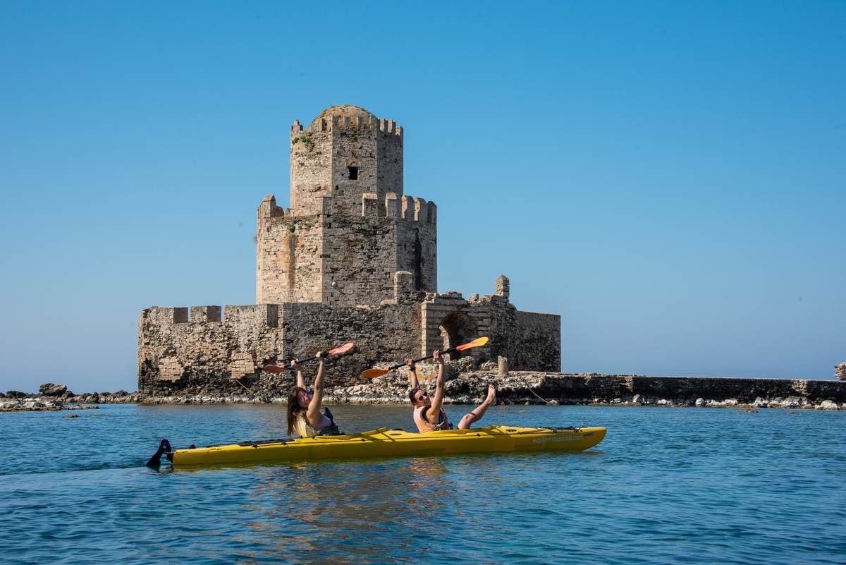 Two women raising paddles in a tandem kayak in front of the Bourtzi tower