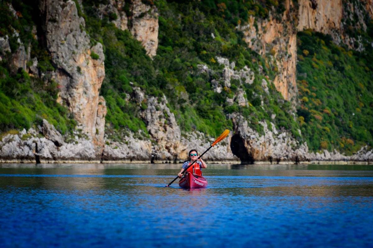 A woman in a red kayak paddling past lush green cliffs and sea caves during a tour in Pylos.