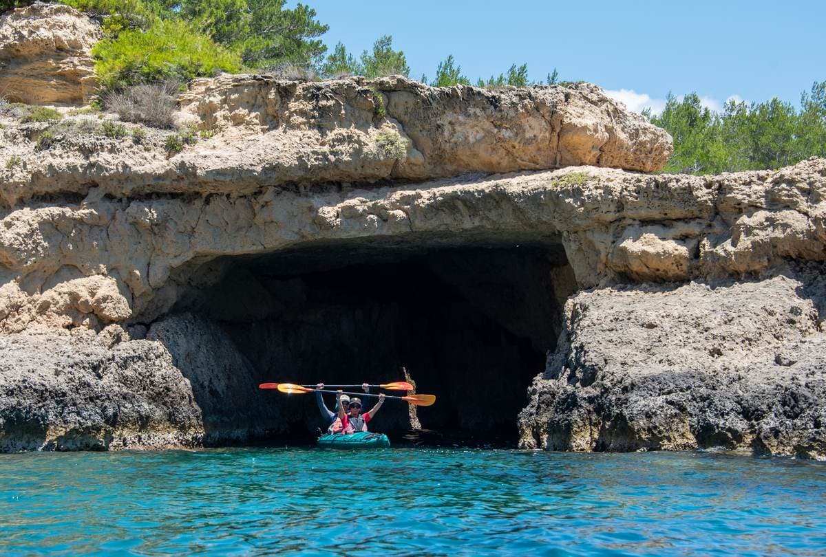 Two people in a sea kayak exploring a large, dark sea cave entrance at the base of a cliff.