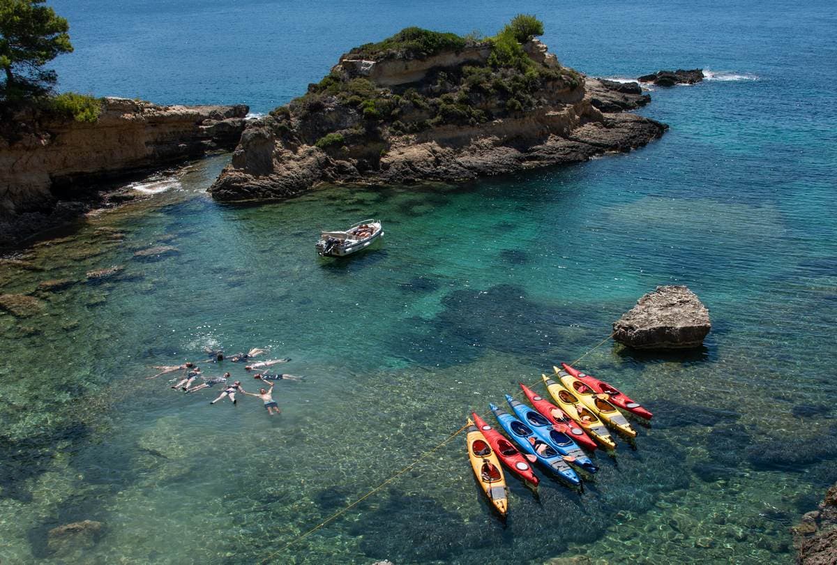 Aerial view of people swimming in a circle and colorful kayaks anchored in the crystal-clear water of Pylos.