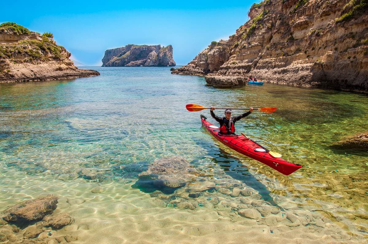 A traveler raising a paddle in triumph on a red kayak in clear, shallow water with a rock arch in the distance.