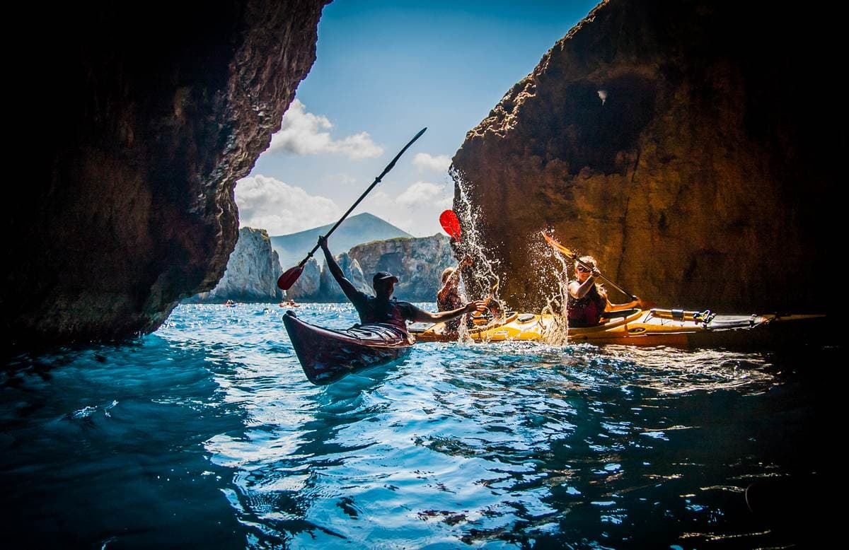 Kayakers inside a sea cave looking out toward the bright blue water and distant hills.