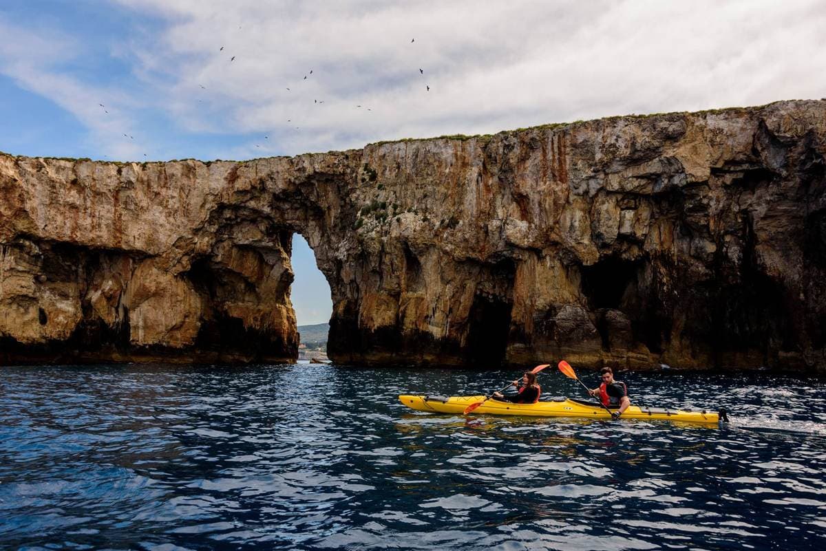 A tandem kayak paddling toward the iconic natural stone arch of Sphacteria Island in Pylos.