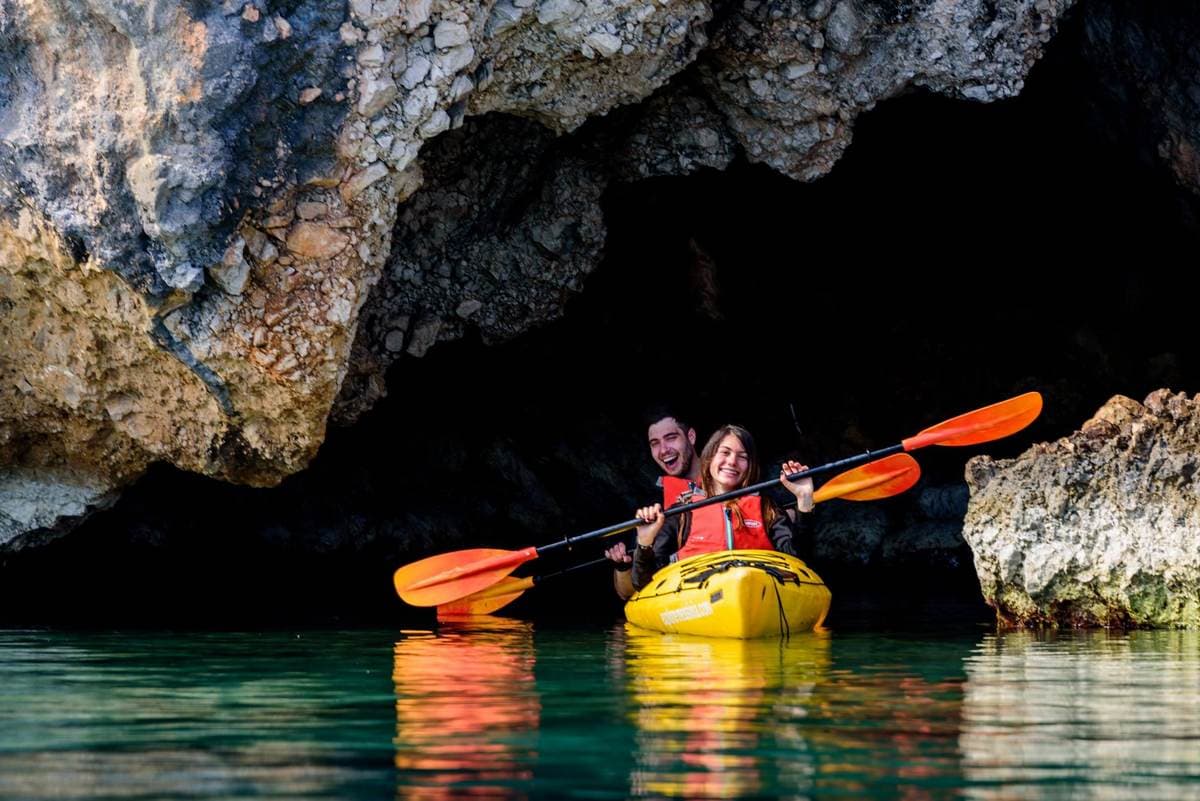 A smiling couple in a yellow tandem kayak entering a dark sea cave during sea kayaking navarino bay.