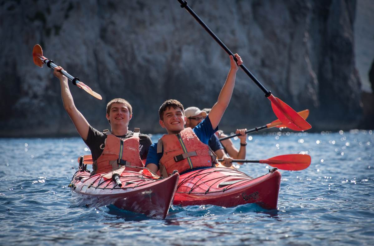 Two cheerful young men raising their paddles in red kayaks during a sea kayaking navarino bay adventure.
