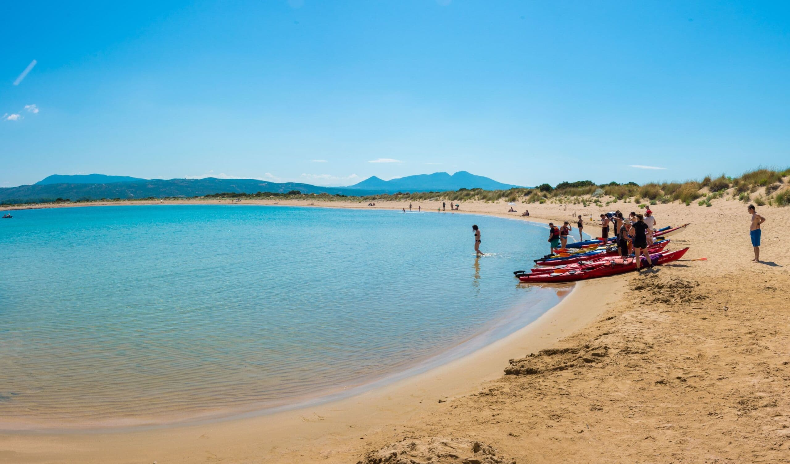 Sea kayaks lined up on the sandy shore of Voidokilia beach with swimmers in the turquoise lagoon