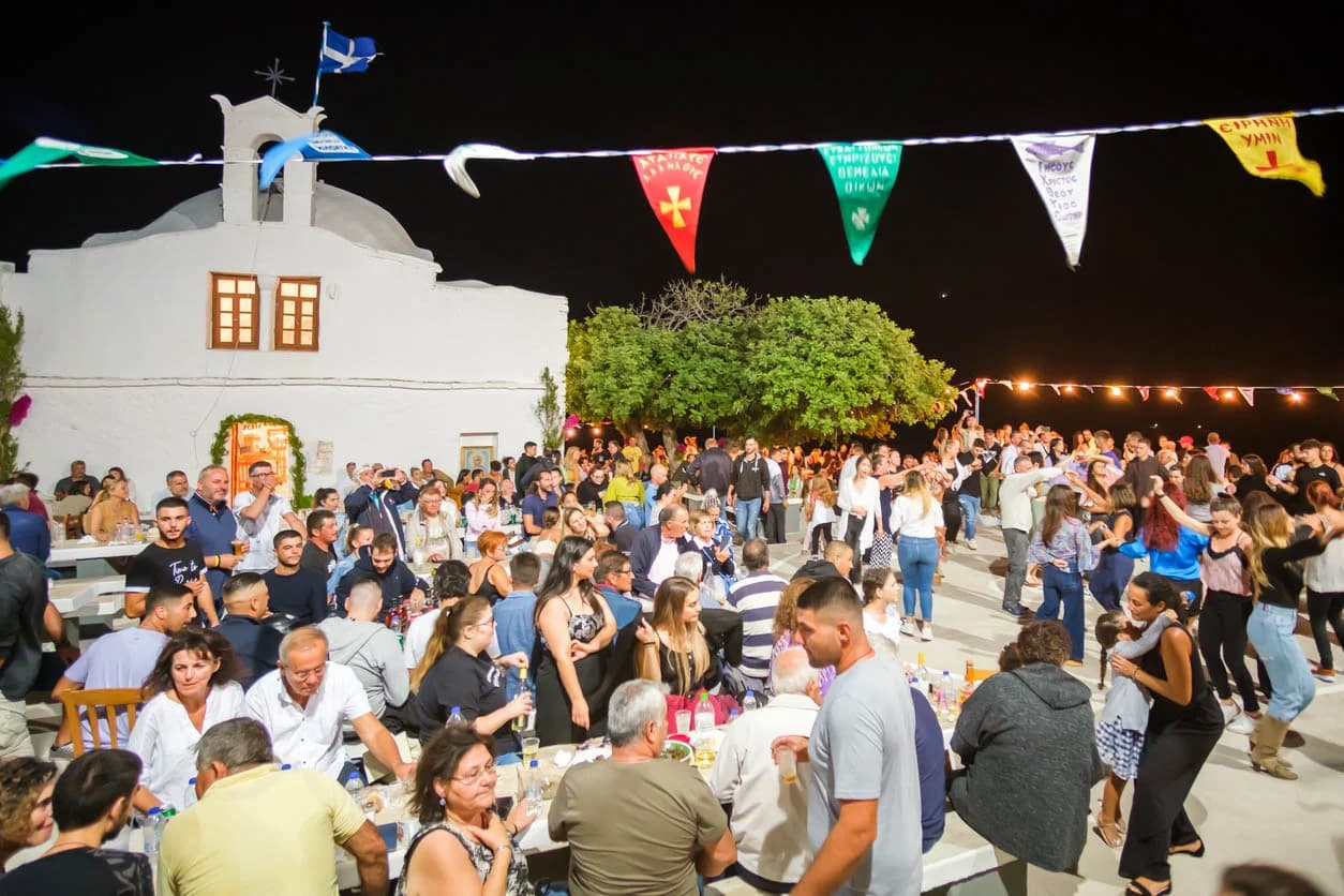 A lively outdoor panigyri in Messinia with colorful flags, a church backdrop, and people enjoying food, drinks, and traditional Greek dancing.