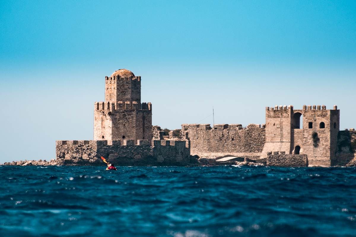 Lone sea kayaker paddling in deep blue water toward the massive medieval walls of Methoni Castle