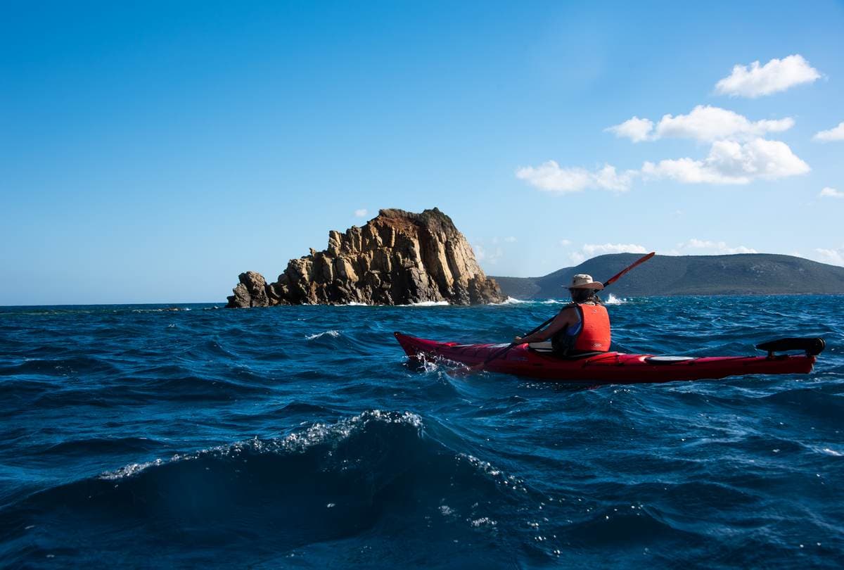A single red kayak navigating choppy blue water near a rocky coastal islet