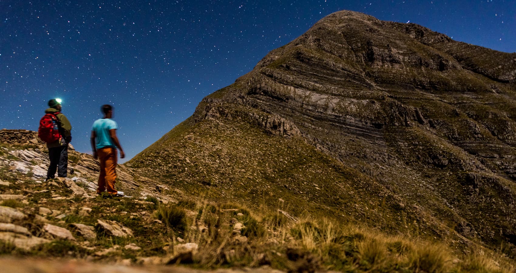 Two hikers with headlamps stand below the rugged Taygetos summit under a starry night sky