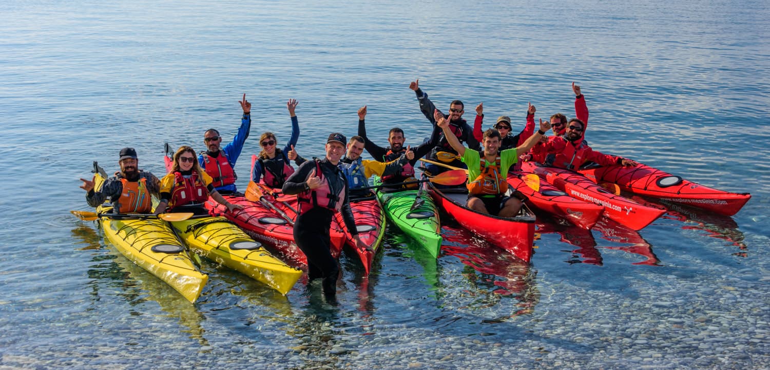 Group of sea kayakers in colorful boats raising arms and cheering in shallow water during a Messinia training day