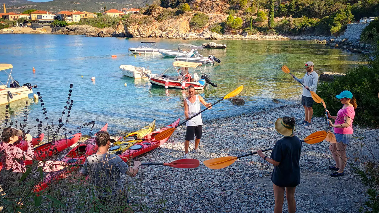 Explore Messinia-A kayaking guide demonstrating safety instructions and paddling techniques to a group of attentive participants, with kayaks and calm waters in the background. A kayaking guide demonstrating safety instructions and paddling techniques to a group of attentive participants, with kayaks and calm waters in the background.