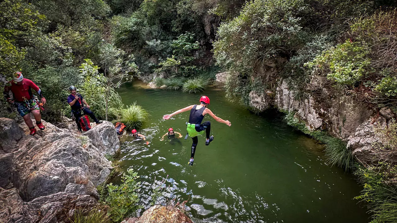 Explore Messinia- Mid-air, leaping into a sparkling turquoise pool framed by rocky terrain at Polylimnio Waterfalls. Explore Messinia- Participant mid-air, leaping into a sparkling turquoise pool framed by rocky terrain and overhanging vegetation at Polylimnio Waterfalls. A snapshot capturing the excitement of family-friendly canyoning in Greece.