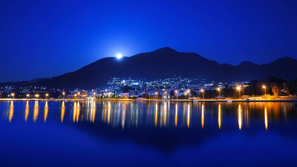 A serene night view of Kyparissia with glowing streetlights A serene night view of Kyparissia with glowing streetlights reflecting on the water and a mountain silhouette under the moonlight.