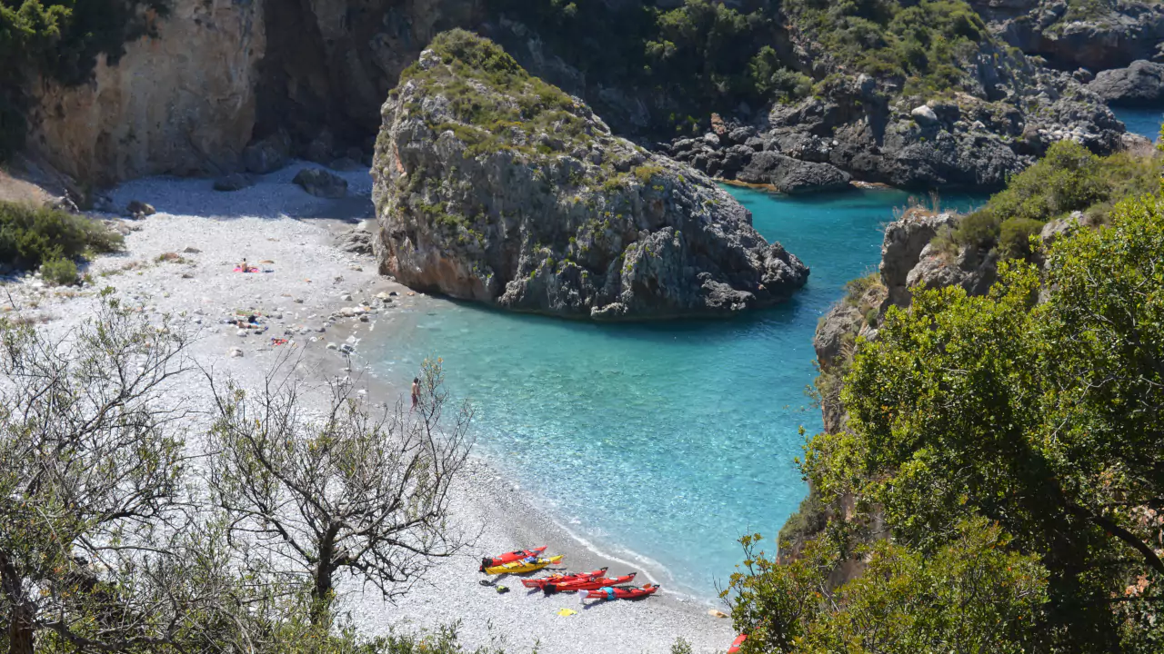 Secluded beach in Mani Greece with colorful kayaks on pebbles, rocky cliffs, and turquoise water.