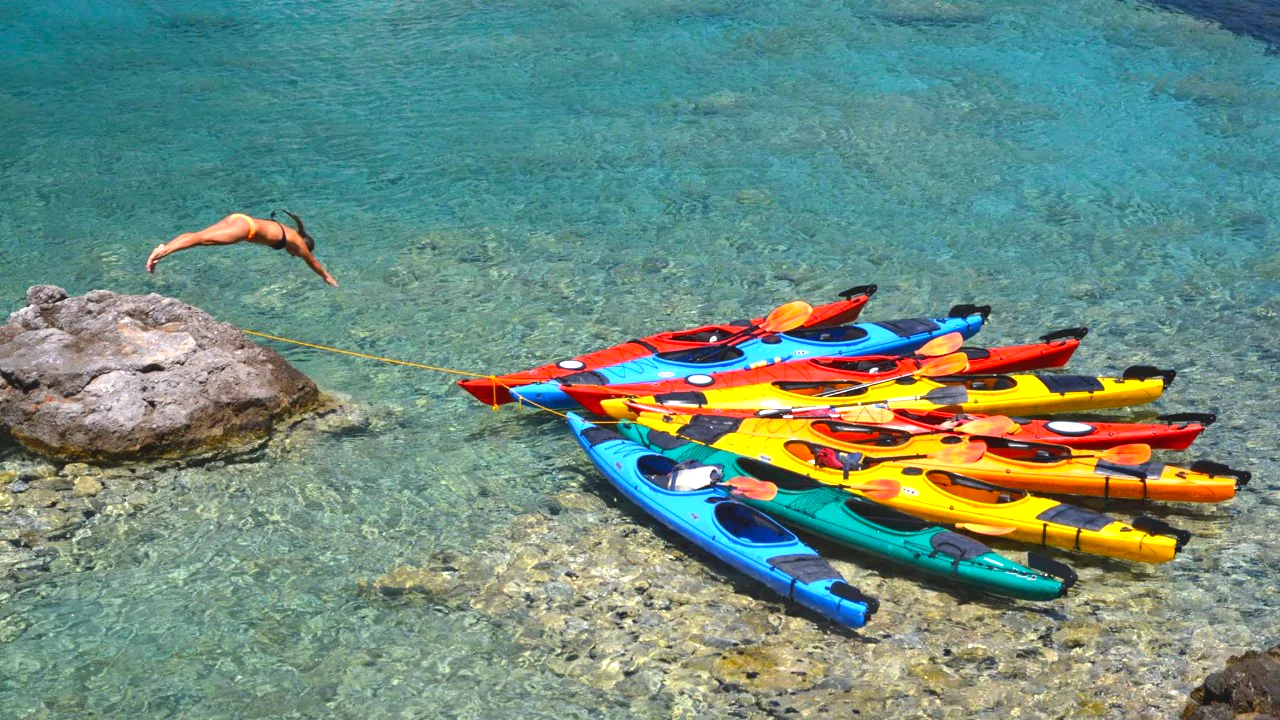 Sea kayaks in turquoise waters near Kardamyli with a person mid-dive from a rock.