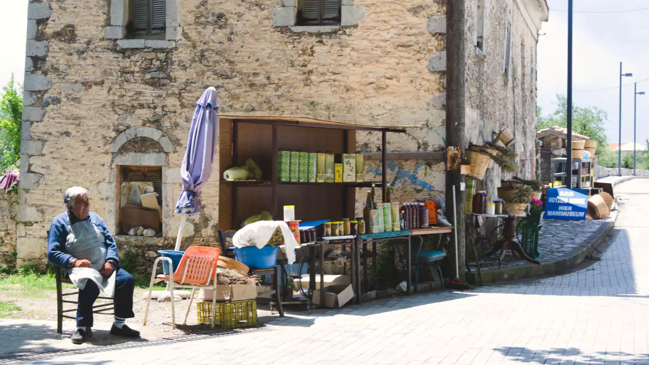 Man at a rustic Mani village market near a stone building showcasing local olive oil and handmade goods.