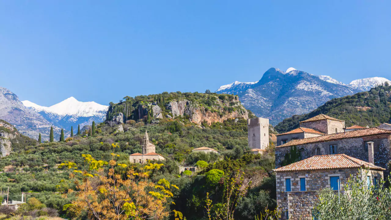 Snow-capped Taygetos Mountains behind green hills, traditional stone buildings with red-tiled roofs, and cypress trees in Mani Greece.